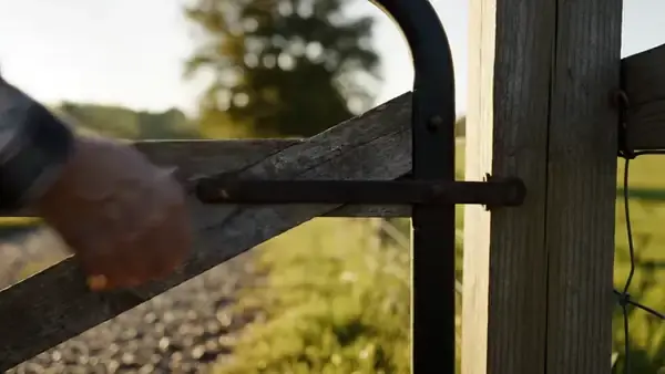 Weathered hand swinging a wooden farm gate closed and dropping the steel latch
