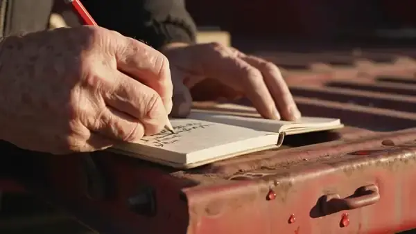 Weathered hand writing a short line with a carpenter's pencil in a pocket notebook on a tailgate